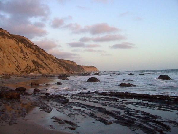 A photo looking out along the coastline where the waves gently roll over the sand and rocks. Cliffs wrap around the water while a few clouds hang above, lurking on an otherwise pleasant day.